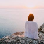 A serene view of a woman sitting on a rocky cliff, gazing at the ocean during sunset. Perfect for relaxation and travel themes.