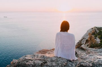 A serene view of a woman sitting on a rocky cliff, gazing at the ocean during sunset. Perfect for relaxation and travel themes.